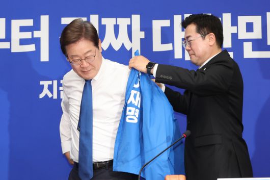 더불어민주당 박찬대 당대표 직무대행 겸 원내대표가 28일 서울 여의도 국회에서 열린 최고위원회의에서 이재명 대선 후보에게 선거운동용 파란색 점퍼를 입혀주고 있다. [사진 출처 = 연합뉴스]