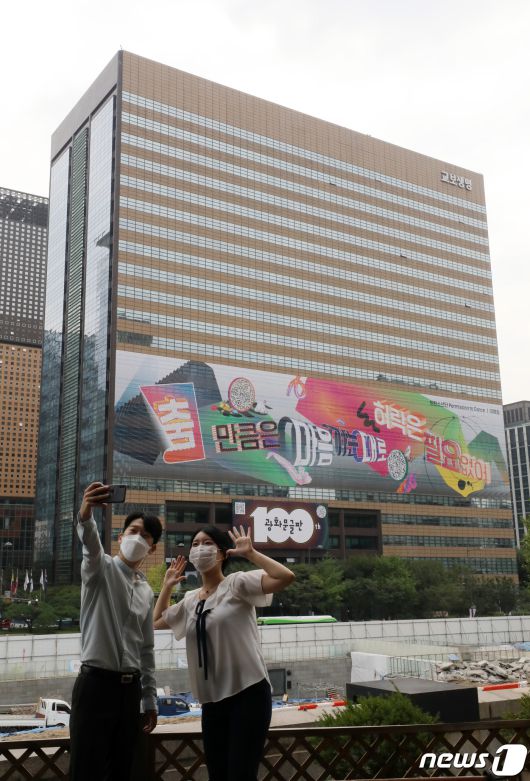 30일 오전 서울 종로구 교보빌딩에 100번째 광화문글판 '춤 만큼은 마음 가는 대로, 허락은 필요 없어'을 배경으로 시민들이 사진을 찍고 있다. 글로벌 아티스트 방탄소년단(BTS)가 100번째 광화문글판을 위해 문안 제작에 참여한 이번 글판은 코로나 펜데믹으로 제약이 늘고 고단한 하루 속에서도 허락받지 않고 자유로움을 느낄 수 있는 것을 찾자는 메시지를 담았다. 2021.8.30/뉴스1 © News1 박세연 기자