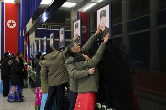4·25문화회관의 실내 추모의 벽에서 희생자 가족들이 초상화를 만지고 있다. 길게 늘어진 인공기와 검은색 벽은 추모의 분위기를 엄숙하게 하기 위한 장치로 읽힌다. (평양 노동신문=뉴스1)