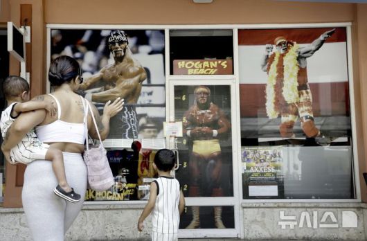 [클리어워터 비치=AP/뉴시스] 24일(현지 시간) 프로레슬러 헐크 호건(본명 테리 진 볼레아)의 사망 소식이 전해진 몇 시간 뒤 미 플로리다주 클리어워터 비치에서 한 여성이 두 아들과 함께 호건의 상점(호건스 비치) 앞을 지나며 그의 사진에 작별 인사를 하고 있다. 미 프로레슬링 전설 '헐크 호건'이 71세를 일기로 별세했다. 사인은 심장마비로 알려졌다. 2025.07.25.