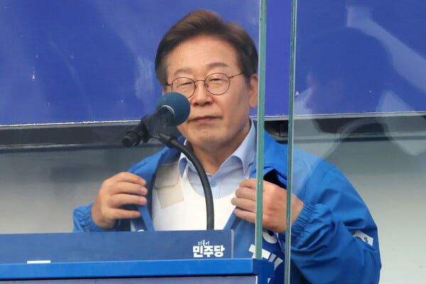 방탄유리막 뒤의 이재명 후보가 21일 인천 계양역 앞 유세에서 선거운동복 지퍼를 내려 방탄조끼를 보여주고 있다. 그는 국민의힘 등에서 방탄 장막을 비판하는 목소리가 나오자 “(지난해)목이 찔린 상대방 정치인을 두고 장난해서야 되겠나”라며 “지금 이 순간에도 반란과 내란이 계속되고 정적 제거 음모가 계속되고 있다”고 했다./뉴시스