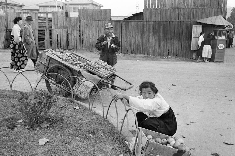 과일 노점상들의 경쟁이 치열하다, 손수레에 과일을 파는 상인이 바로 옆에 사과 한 박스를 그대로 놓고 파는 여자를 노려보자 여자는 고개를 돌리고 있다. 손수레 상인은 무섭게 노려보며 칼을 들고 있지만 실은 과일을 깎아 한입 물고 있다. 살아 남기 위해 치열하게 경쟁했던 시대, 하지만 볼 수록 웃음이 난다. 이들 뒤로 걸어가는 멋지게 차려입은 신사와 부녀자가 그당시 빈부 격차도 느끼게 한다. /©한영수_서울 1956-1963