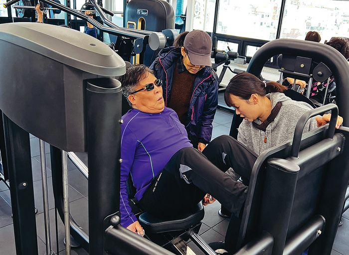 아내와 함께 헬스장을 찾은 시각장애인 어르신이 헬스장 운영요원(오른쪽)의 안내에 따라 운동기구(레그프레스)에서 자세를 잡고 있다.