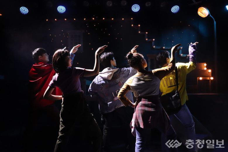 어둡게 연출된 무대 위에서 다섯 명의 배우가 조명 방향을 향해 팔을 들어 올린 채 퍼포먼스를 펼치고 있다. 후방에는 은은한 조명과 계단 형태의 세트가 배치돼 장면의 분위기를 극적으로 만들며, 젊은 배우들의 활기와 움직임이 무대 전체에 생동감을 더한다