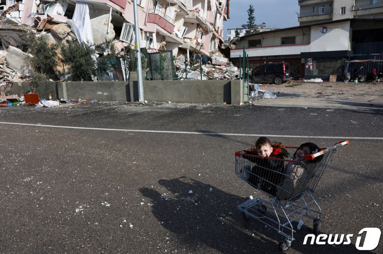 7일(현지시간) 튀르키예 하떠이 한 주택가에 두 꼬마가 시장 카트 속에 쪼그려 앉아 있다. 아이들의 뒤로는 강진으로 무너진 아파트와 잔해가 뒤죽박죽 섞여 있다. ⓒ 로이터=뉴스1 ⓒ News1 권진영 기자