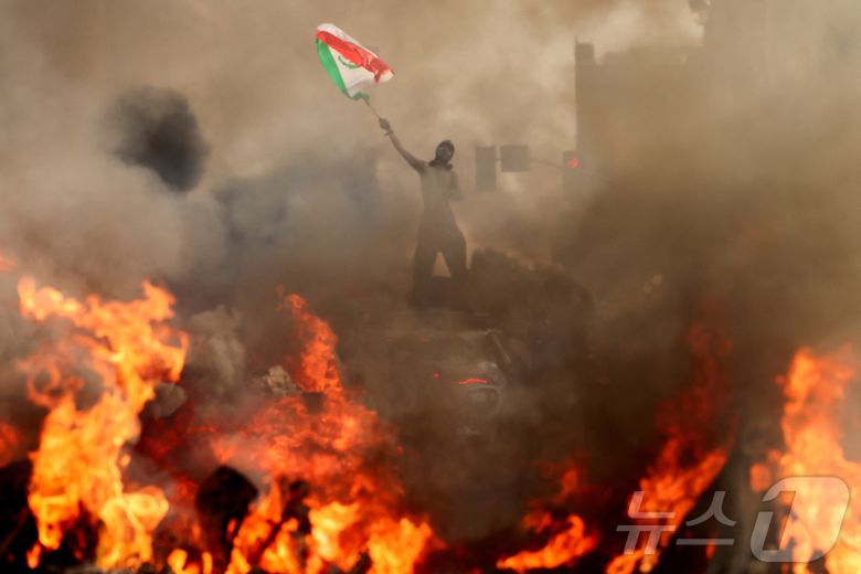 한 남자가 로스앤젤레스 시청 근처에서 연방 이민 단속에 반대하는 시위 중에 연기가 나고 불길이 치솟는 차량 옆에서 멕시코 국기를 흔들고 있다. 2025.6.8 ⓒ 로이터=뉴스1 ⓒ News1 강민경 기자