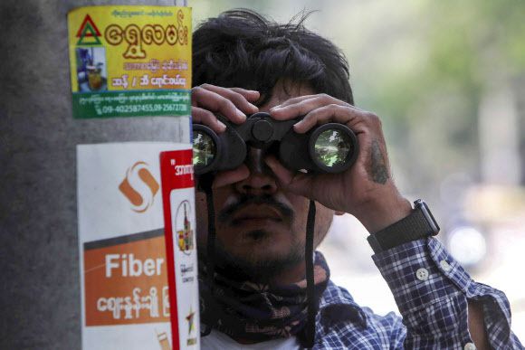 지난 18일 미얀마 만달레이에서 한 시위 참가 남성이 미얀마 군인들이 불도저로 반 쿠데타 시위대가 설치했던 모래주머니 방호벽을 철거하는 모습을 쌍안경으로 지켜보고 있다. 만달레이=AP 뉴시스